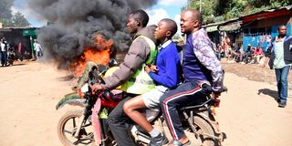 A student (centre) is ferried on a motorcycle as it passes next to a bonfire in Kibera, Nairobi